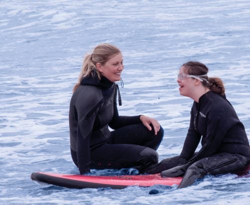 Emily Clapham sitting on a surf board with Tessa Eagan at Narragansett Beach.