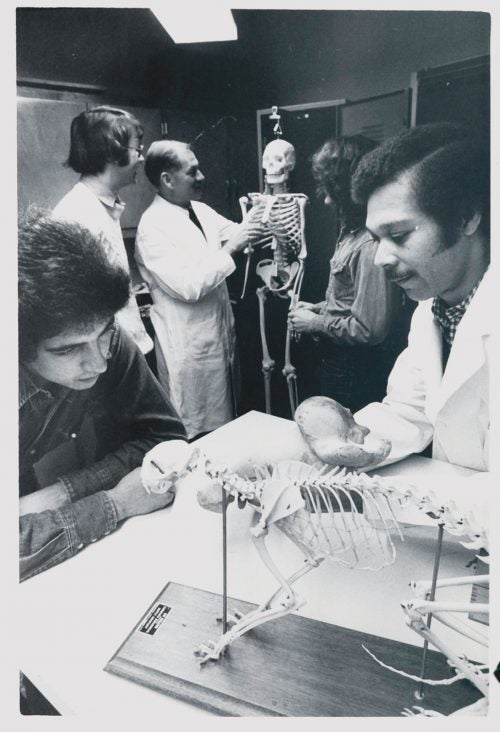 An old black and white photo of students and professors in a lab studying animal skeletons
