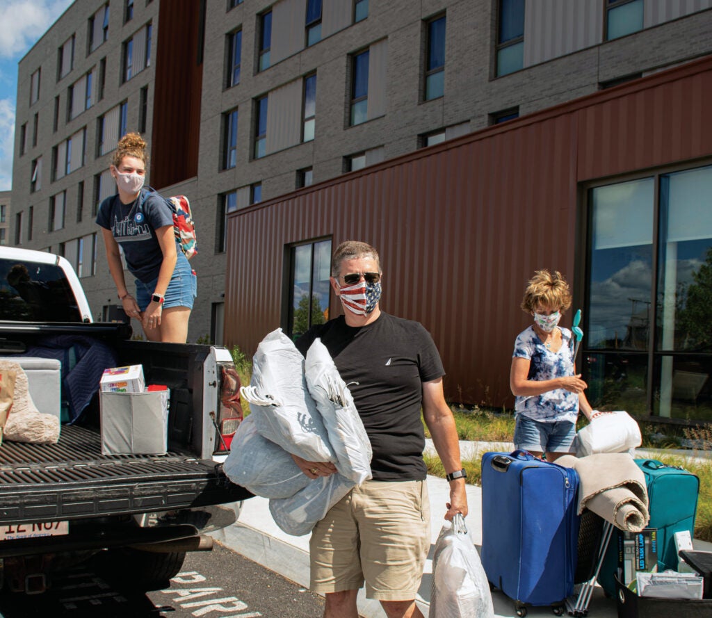 Three people wearing masks unloading a pick-up truck, parked in front of a dorm