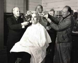A historical black and white image of a person getting his hair clipped as two others look on.