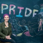 URI Doctoral student and Fulbright recipient Meg Jones seated before a colorful chalk drawing with the word PRIDE in rainbow letters