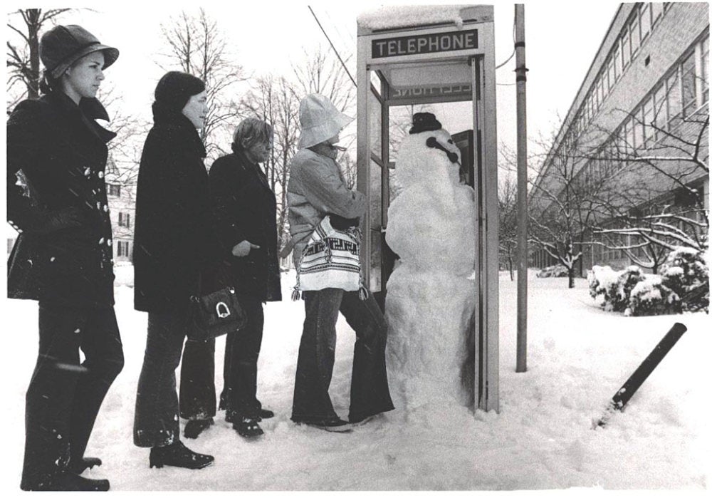 A black and white picture of students during the winter waiting outside the telephone booth occupied by a snowman.