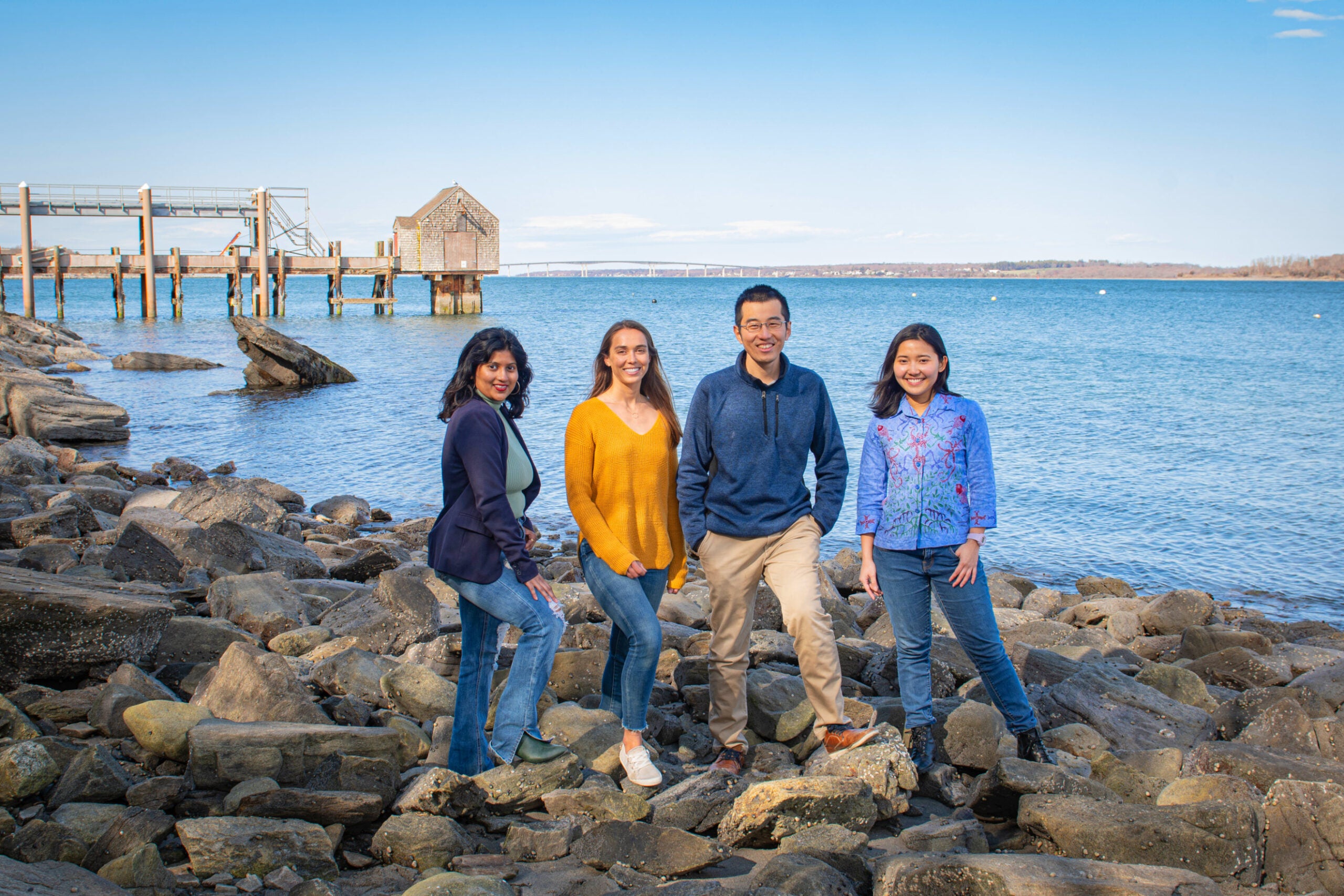 Hacking4Oceans graduate students at the bay campus on the rocks with the dock in the background: Monica Rao; Christine De Silva; Erfa Fachroni; Xiaozhuo Wei