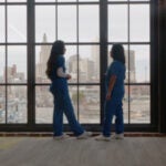 Two students wearing scrubs, looking out a large window at a cityscape