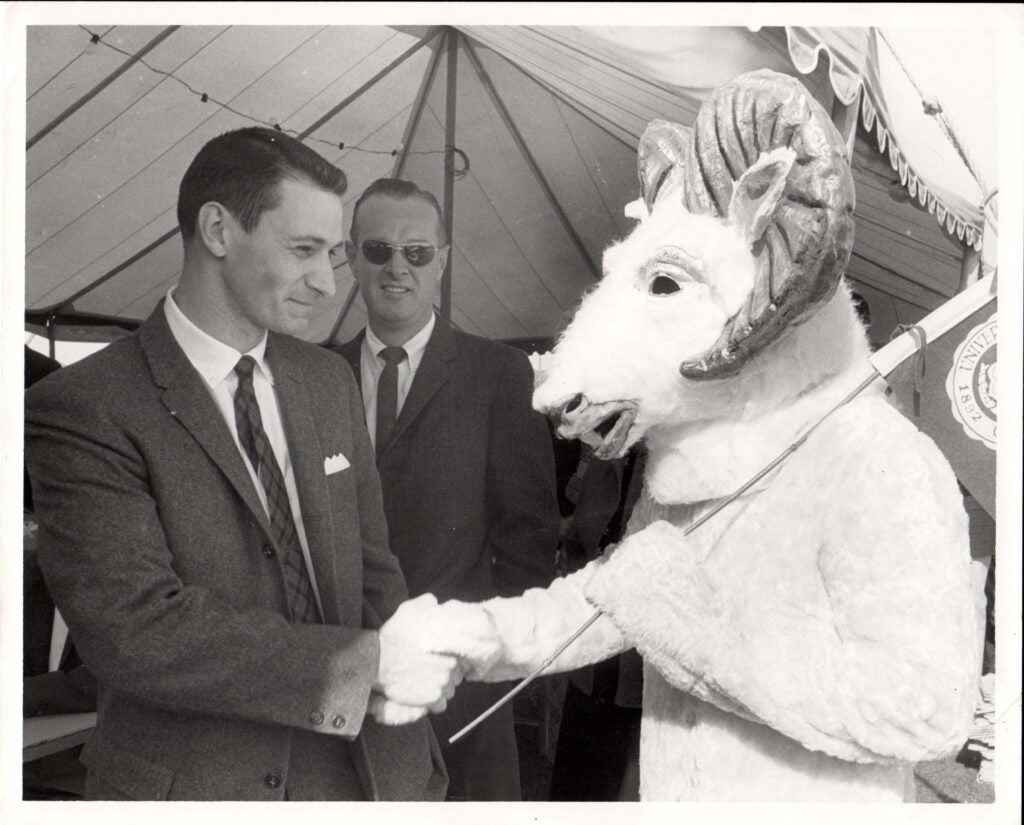 A black and white image of a man in a business suit shaking hands with the URI mascot