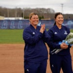 URI coaching colleagues Whitney Goldstein and Tori Constantin stand smiling in a softball field, Constantin holds flowers