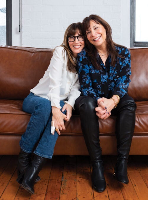 Sisters Abby Rodman ’84 and Julie Howard ’86 hosts of Sisters Cracking Up, a podcast focused on midlife seated with their heads leaning together.