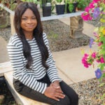 Tolani Olagundoye is a clinical assistant pro- fessor of leadership in the College of the Environment and Life Sciences and a Coastal Institute fellow, seated, surrounded by blooms in the URI greenhouse