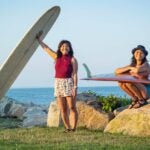 Graduate students and surfers Juliet “Satya” Sullivan and Lauren Zane pose with their surfboards on a rocky shoreline with the ocean behind them at sunset