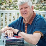 Author Julien Ayotte '63 sitting outdoors in a short sleeve t-shirt with a stack of his books
