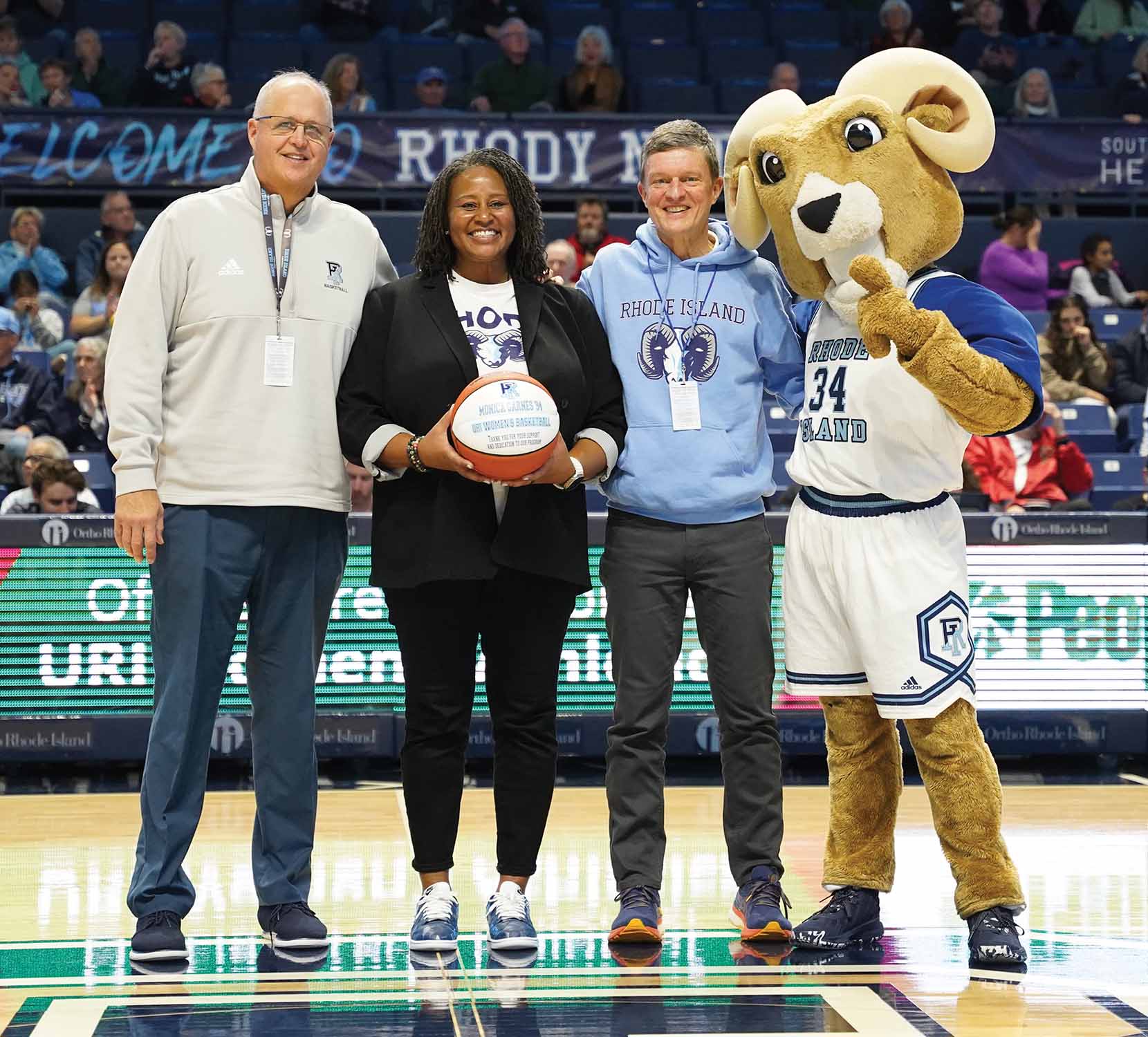 Monica Garnes '94 on the URI basketball court with President Parlange and Rhodey Ram. Monica is holding a URI branded basketball.