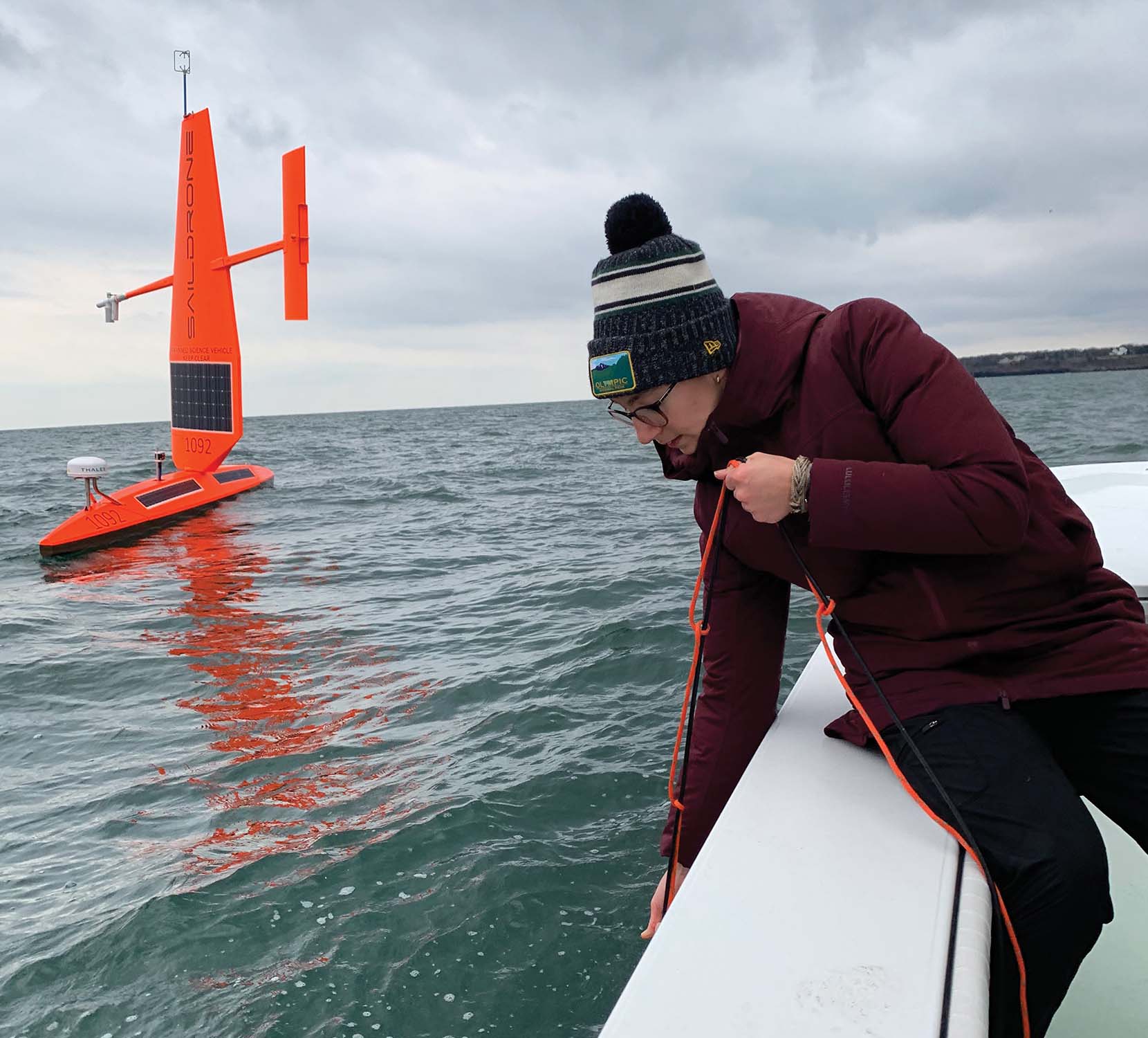 Sarah Nickford '23 in winter gear, leaning over the side of a boat holding a rope with a Saildrone in the background