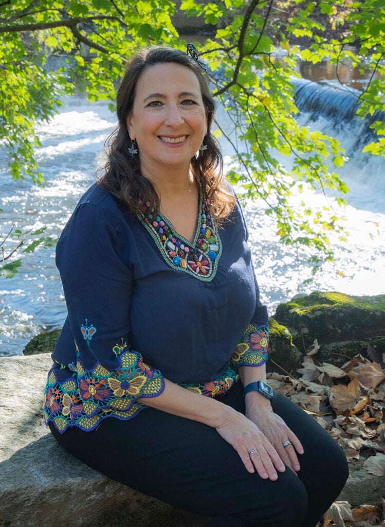Alicia Lehrer, M.S. '93, Executive Director, Woonasquatucket River Watershed Council. Alicia wears a dark blue shirt and sits in front of the Woonasquatucket river with green foliage behind her