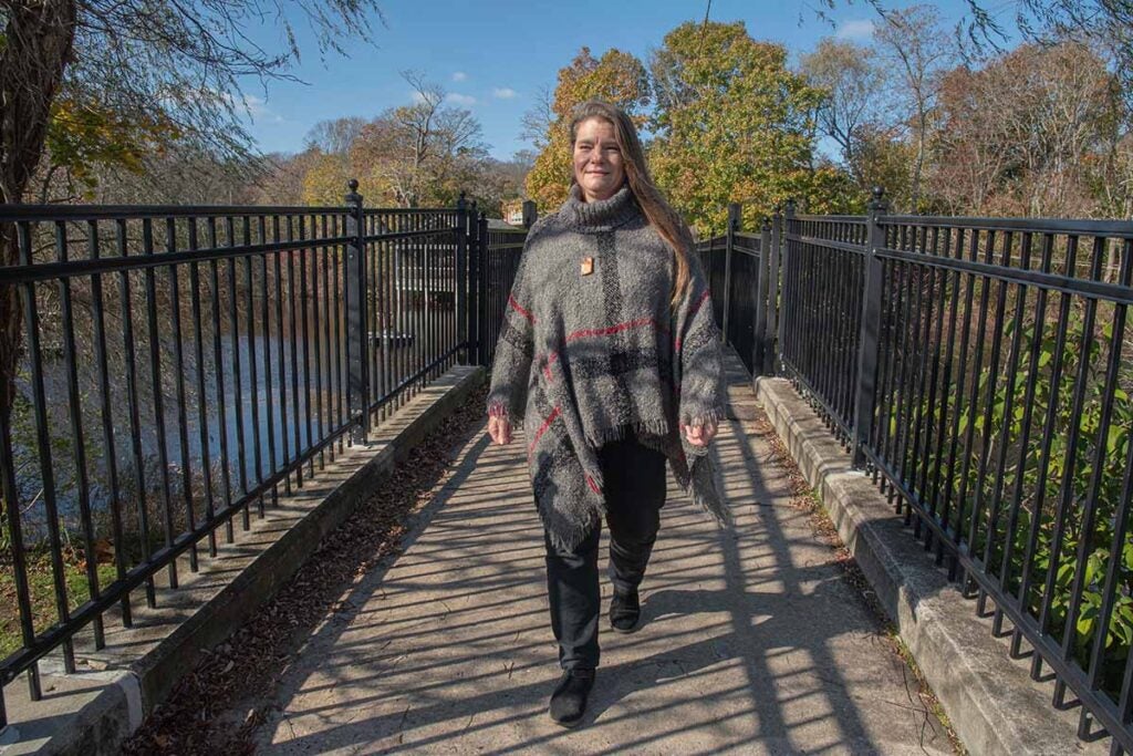 Elizabeth Herron '88, M.A. '04, Director of URI's Watershed Watch. Elizabeth wears a long sleeve sweater and jeans and walks towards the camera with the Saugatucket River behind her