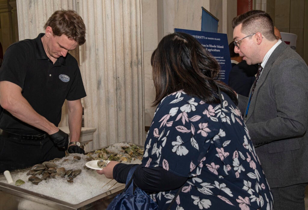 A man shucks oysters behind a table while two other individuals look on
