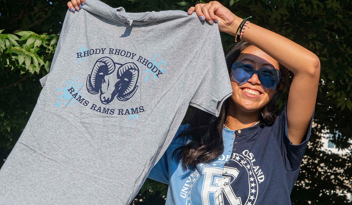 female student in a RI Rams shirt holding up a Rhody T shirt and smiling