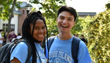 a female and male student wearing URI t shirts smiling widely near the Union on a bright sunny day